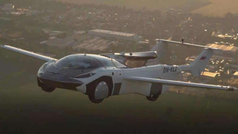 Kanatlarını açmış fütüristik bir uçan araba, gün ışığında tarlalar ve binalardan oluşan bir manzaranın üzerinde süzülüyor. Araç, aerodinamik tasarımı ve görünür kayıt numarasıyla hem bir otomobilin hem de bir uçağın özelliklerini bir araya getiriyor.