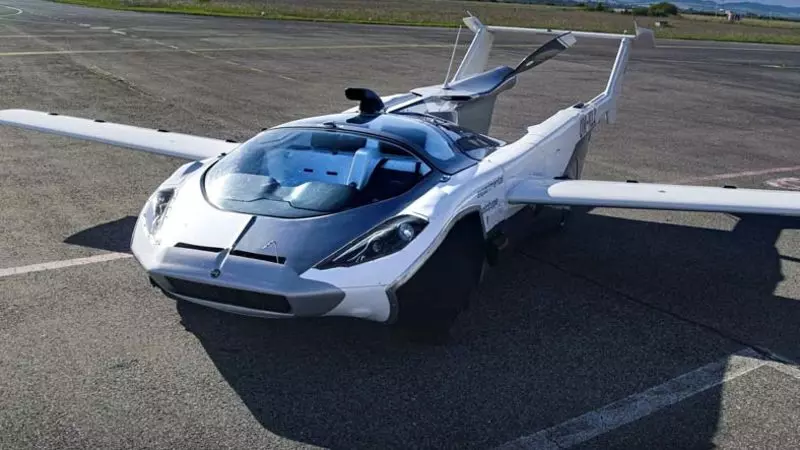 Kanatlarını açmış gümüş renkli bir uçan araba, fütüristik tasarımını ve aerodinamik özelliklerini sergileyerek boş bir piste park edilmiştir.